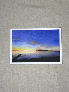 A postcard featuring "Morning Sunrise," an original watercolor painting of a lake at sunrise with a dock on the left and an island in the distance, displayed on a beige fabric surface.
